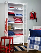White display cabinet with open doors and the pillows and blankets inside, in front red metal step stool next to a wicker chair
