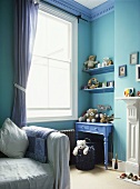 A detail of a child's blue bedroom with a painted table, shelves, gingham curtains, soft toys, covered sofa