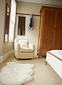 A detail of a neutral traditional bedroom with a free standing wardrobe, upholstered armchair, furry rug