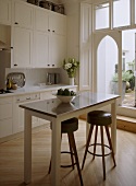 A detail of a modern, country kitchen with dining area, painted units, table, pair of stools, wood floor, door out to garden