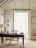 A traditional study room decorated in neutral colours, pattern wallpaper, beamed ceiling, wooden table, chair, lamp, natural flooring, French windows,