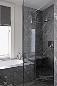 Shower unit with glass door next to bathtub below window with grey roman blind in contemporary bathroom