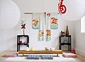 Papierhängelampen und Sitzpolster vor Bodentisch im japanischen Wohnzimmer mit weiss getäfelter Wand und hängendem Kimono
