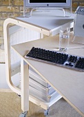 A detail of a modern home office showing a desk, computer screen, keyboard, portable trolley storage unit on wheels