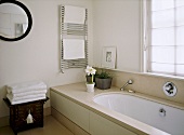A detail of a modern, neutral en suite bathroom showing bath, chrome towel rail radiator, chest