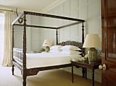 A traditional bedroom with painted panelling, carved wooden four poster bed, bedside table, lamps