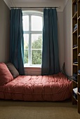 A double bed with a pink plaid cover next to a window with blue curtains