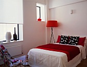A floor lamp with a red shade next to a double bed with black and white cushions and a red quilt