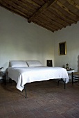 A high double bed with white bedclothes in a bedroom with a terracotta floor and a wood beam ceiling