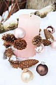 A pink candle with pine cones and Christmas baubles in the snow
