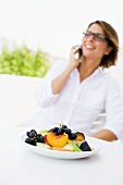 Plate of fruit, woman using mobile phone in background
