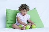 A little girl playing with green apples on a sofa