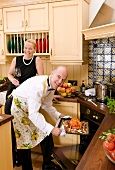 Man putting duck into oven, woman behind with glass of red wine
