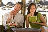 Father eating watermelon, daughter drinking coconut milk on beach