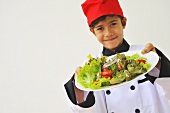 A boy dressed as a chef holding a plate of salad