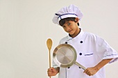 A boy dressed as a chef holding a pan and a wooden spoon