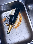 An empty champagne bottle, cutlery and leftover food in a sink