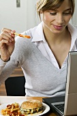 Woman eating hamburger & chips while working at computer