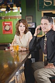 Frau steht missbilligend neben telefonierendem Mann im Pub