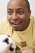Young man with football and chocolate bar watching TV