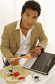 Asian man eating sushi while sitting at laptop