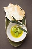 Green olives on twig in bowl of olive oil, crackers