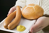 Person holding frankfurters, roll & mustard on paper plate