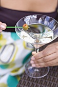 Woman holding green olive on cocktail stick over Martini glass