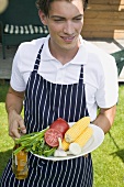 Young man in apron holding vegetables & beer at a barbecue