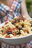 Woman holding large dish of pasta salad with olives & tomatoes