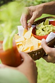 Hands holding basket of guacamole & chips and tomato drink