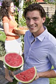 Young man holding watermelon, woman with iced tea in background