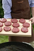 Man holding raw burgers on chopping board over barbecue