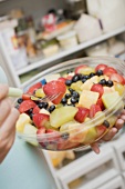 Woman eating fruit salad out of plastic bowl in front of refrigerator