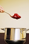 Hand holding a spoonful of red berry compote over a pan