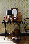 Kleiner Tisch mit Holzskulpturen, einer Tischlampe, holzgerahmtem Spiegel und antikem Vogelkäfig vor Wandtapete mit floralem Muster