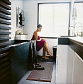 Junge Frau in ein violettes Badetuch gewickelt, in modernem Badezimmer sitzend