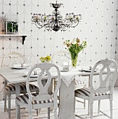 Gedeckter, rustikaler Esstisch in Weiß mit antiken Stühlen, einer Spitzendecke und einem Frühlingsblumenstrauss