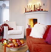 Wohnzimmer mit brennendem Feuer im Kamin, Kerzendeko auf dem Kaminsims und einem plüschigen roten Sofa mit weißem Fellkissen