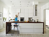 Junge Frau in moderner Küche mit großem Küchenblock und zylinderförmigem Dunstabzug aus Edelstahl