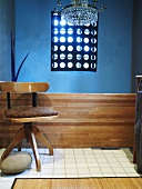 Drehstuhl aus Holz mit braunem Sitzpolster vor blauer Wand; darüber ein Kristallleuchter