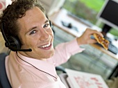Junger Mann mit Kopfhörer und einem Stück Pizza im Büro