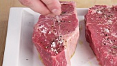 New York strip steaks being seasoned with salt