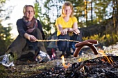Two women cooking sausages on a campfire