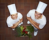 Two chefs with their arms folded beside crate of vegetables