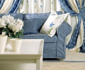Blaues britisches Sofa davor ein weißer Tisch, Gardinen in blau/weiß.