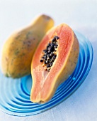 Halbierte Papaya mit Kernen neben ganzer Frucht auf blauem Teller