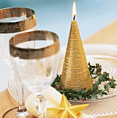 Goldene Kerze in Kegelform steht auf Glasteller in einem Kranz aus Ilex