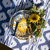 Keramikteller mit Sonnenmotiv, blaues Weinglas, Sonnenblume