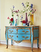 Verschiedenfarbige Glasvasen mit Blumen auf blauer Rokoko Kommode mit vergoldeten Schnitzereien
